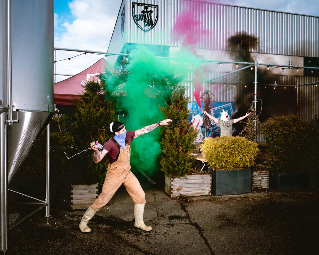 Photo promo pour la nouvelle bière éphémère Chaosphère - new england ipa Sainte Cru brasseur devant la brasserie Sainte Cru à Colmar représentant le graffiti de Banksy "Flower Thrower" avec des fumigènes de couleurs encouragé par une personne avec un masque de licorne qui en tient un rose